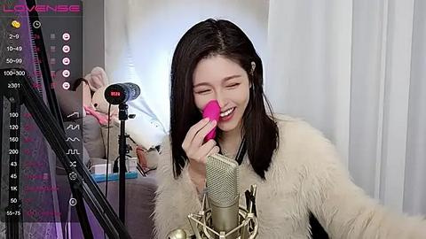 Media: A video of a smiling Asian woman with long black hair, wearing a beige fur coat, holding a pink makeup brush near a microphone on a grey sofa.