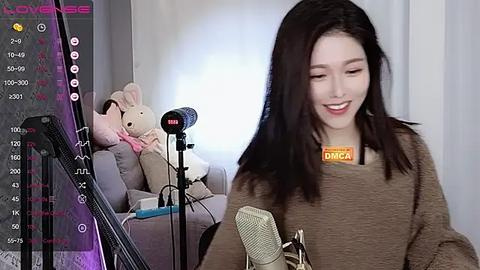 Media: Video of a young, fair-skinned Asian woman with long black hair, smiling, wearing a brown sweater, microphone, and plush toys in a cozy room.