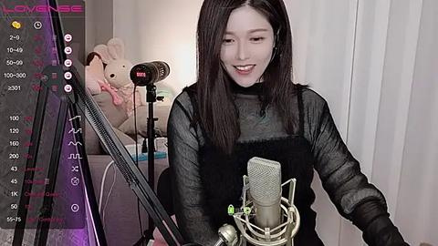 Media: Video of a smiling, slender Asian woman with straight black hair, wearing a black top and apron, singing into a microphone in a dimly lit room with a stuffed animal and recording equipment.