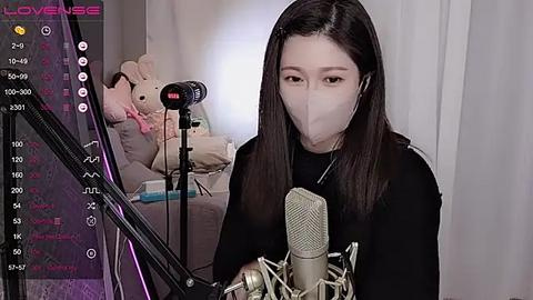 Media: Video of an East Asian woman with long black hair and a mask, singing into a microphone, surrounded by a cozy, dimly lit room with a plush teddy bear.