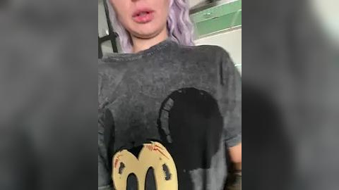 Media: A video of a person with light purple hair, wearing a gray graphic tee with a black-and-yellow cartoon face design. The background features a green door and white wall.