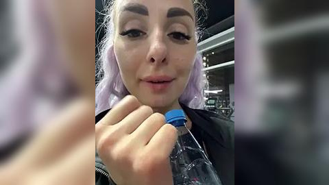 Media: Video of a fair-skinned woman with light purple hair, wearing black clothing, holding a plastic water bottle, taken indoors with a blurred background.