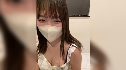 Media: Video of an Asian woman with straight, shoulder-length brown hair, wearing a white face mask and a floral-patterned dress, indoors with a TV in the background.