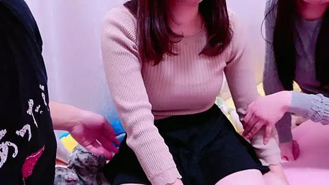 Media: Video of a young woman with medium skin tone, shoulder-length brown hair, wearing a ribbed beige sweater and black skirt, sitting on a pink bed, surrounded by others, in a room with pastel-colored walls.