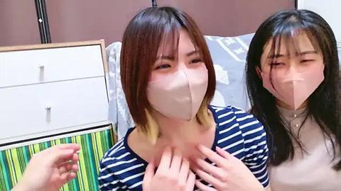 Media: Video of two East Asian women with short brown hair, wearing beige masks, holding hands. They sit on a bed with striped bedding, next to a white dresser with a green handle.