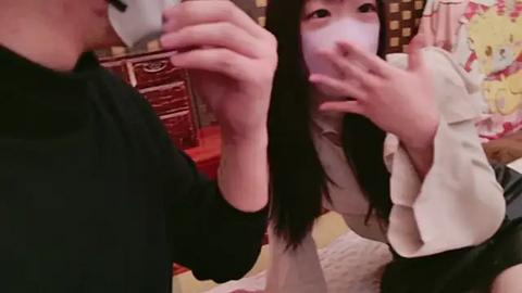 Media: Video of an East Asian woman with long black hair, wearing a white face mask and a beige blouse, taking a selfie, while a man in a black shirt holds a camera.