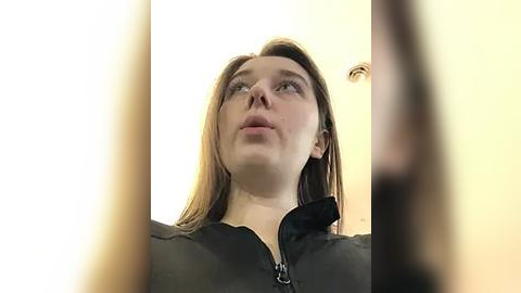 Media: Video of a young Caucasian woman with straight, light brown hair, wearing a black zip-up hoodie, looking upwards with a thoughtful expression. The background is a blurred, light-colored indoor setting.