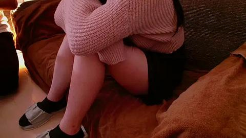 Media: Video of a person sitting on a bed, wearing a pink knitted sweater and black skirt, with their knees pulled up, black socks, and gray sneakers.