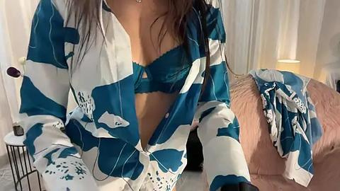 Media: Video of a woman in a teal and white abstract-patterned kimono, revealing a teal lace bra, sitting on a bed with a pink blanket.