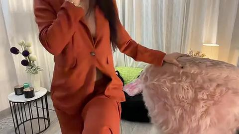 Media: Video of a woman with long brown hair in an orange blazer and matching pants, touching a soft pink fur throw on a black couch. Background features white curtains, a modern table lamp, and a green pillow.