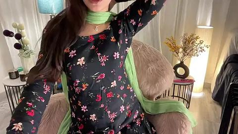 Media: Video of a woman with long brown hair, wearing a black floral dress with green scarf, sitting on a beige chair in a modern, softly lit room with white curtains and decorative vases.