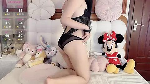 Media: A video of a slender, fair-skinned woman with long black hair, kneeling on a bed in black lingerie. The room features a white headboard, plush toys, and a Minnie Mouse plush.