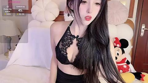 Media: A video of a young Asian woman with long black hair and fair skin, wearing a black lace bralette, sitting on a bed with Disney-themed pillows.