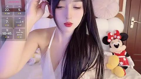 Media: Video of an Asian woman with long black hair, red lips, and white spaghetti straps, adjusting her hair in a bedroom with Minnie Mouse plush toys.