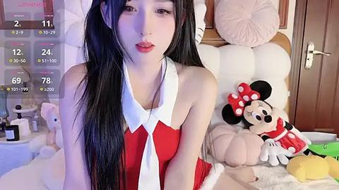 Media: Video of an Asian woman with long black hair, wearing a red Santa dress, sitting on a bed with plush toys, including Mickey Mouse.