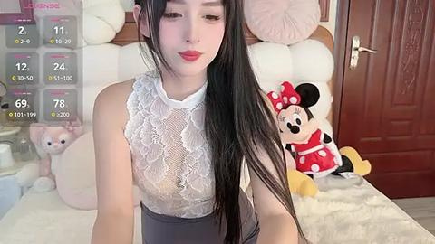 Media: A video of a young Asian woman with long black hair, fair skin, and a slender figure, wearing a white lace top and high-waisted pants, sitting on a bed with a plush Minnie Mouse doll beside her.