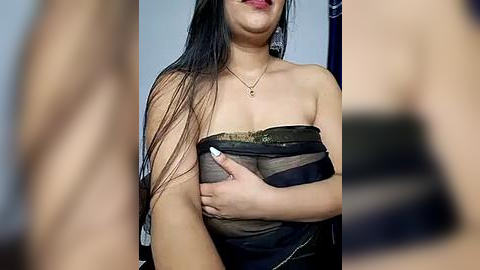 Media: Video of a topless woman with long black hair, holding a black purse against her chest, wearing a gold necklace. The blurred background features a dark curtain and a white wall.