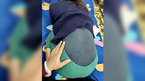 Media: Video of a person lying on their stomach on a colorful patterned blanket, wearing a dark blue sweater and teal pants. Another person\u2019s hand rests on their back, indicating support or comfort.