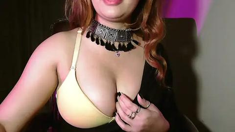 Media: A video of a light-skinned woman with wavy auburn hair, wearing a yellow bra and a black choker with dangling beads, posing indoors under dim lighting.