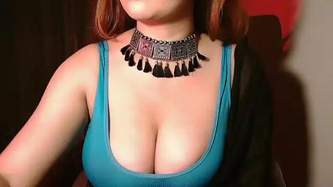 Media: Video of a woman with light skin and shoulder-length auburn hair, wearing a turquoise tank top revealing ample cleavage, and a silver, beaded necklace with tassel accents. Background is dark with a hint of red.