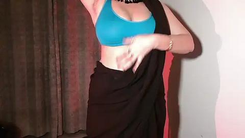 Media: Video of a woman with fair skin and light blue bra, black skirt, and black top, partially lifting skirt, standing in front of brown curtains.