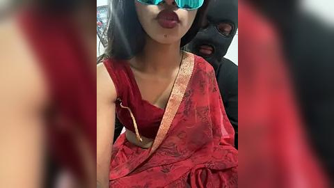 Media: Video of a woman with dark hair, fair skin, and red lipstick, wearing a red saree and black sunglasses, sitting in a car with a masked driver, blurred background.