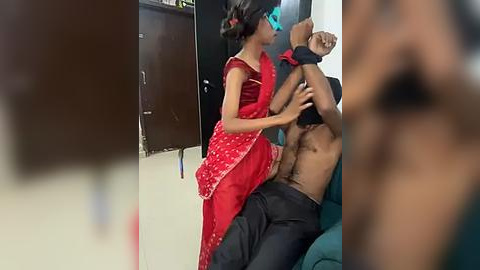 Media: Video of a South Asian woman in a red sari, restraining a shirtless man in black pants, in a domestic setting with a blurred background.