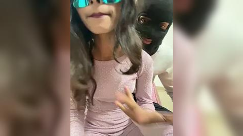 Media: Video of a young girl with dark hair and pink dress, wearing blue sunglasses, and a black dog wearing a black mask, both indoors.