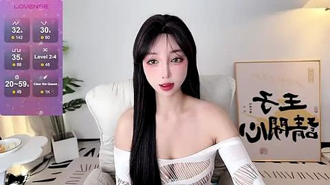 Media: A video of a young East Asian woman with long black hair, fair skin, and red lipstick, sitting on a bed with white pillows and a framed painting in the background.