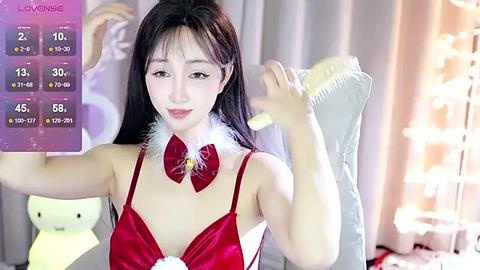 Media: Video of a young Asian woman with long black hair, wearing a red, low-cut, satin lingerie set with a white bow, sitting in a plush chair in a softly lit room.