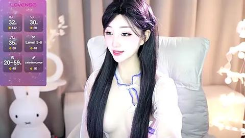 Media: A video of a young Asian woman with long black hair, fair skin, and a slender build, wearing a white blouse. She sits on a cushioned chair against a softly lit background with white curtains and a bunny figurine.