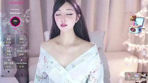 Media: A video of an Asian woman with long black hair, closed eyes, and pale skin, wearing a white floral kimono. The background features a soft-focus, beige-colored room with a white bed.