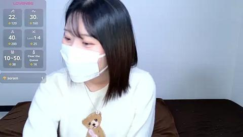 Media: Video of an East Asian woman with straight black hair, wearing a white mask and a white shirt with a teddy bear graphic, indoors against a plain white wall, with a COVID-19 symptom tracker overlay.