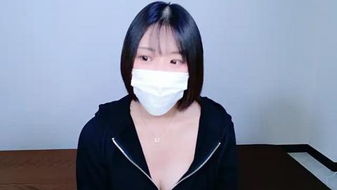 Media: Video of an Asian woman with shoulder-length black hair, wearing a white face mask, black hoodie, and necklace, standing against a plain white wall, indoors.