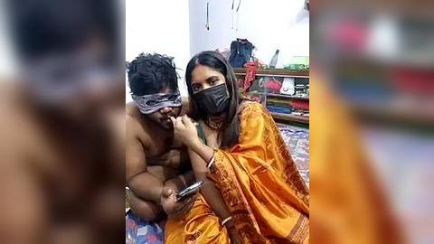 Media: Video of a shirtless man with a blindfold, sitting next to a woman in an orange saree, both holding smartphones in a cluttered room with colorful fabrics on shelves.