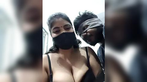 Media: Video of a South Asian woman with a medium complexion, wearing a black mask and revealing black lingerie, standing beside a man with a bandage over his eyes. The background is blurred.