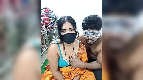 Media: Video of a man with a black mask and orange cloth, hugging a woman in a blue bra and orange sari, against a blurred background.