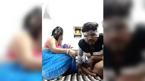 Media: Video of a South Asian woman in a bright blue saree and a man in a striped shirt, both wearing headbands, kneeling on a striped carpet in a dimly lit room with a small yellow lamp.