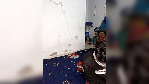 Media: Video of a man with short dark hair, wearing a black jacket, kneeling in a small room with a white wall and a blue bed with cartoon characters, surrounded by clutter and a dangling wire.