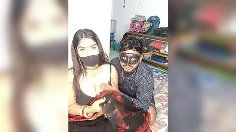 Media: A video of two masked people, a woman with long black hair and a man, seated on a patterned carpet, surrounded by colorful bags in a cluttered room.