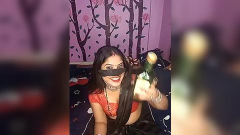 Media: Video of a smiling woman in a red saree, black blindfold, and choker, holding a camera, against a purple wall with floral patterns.