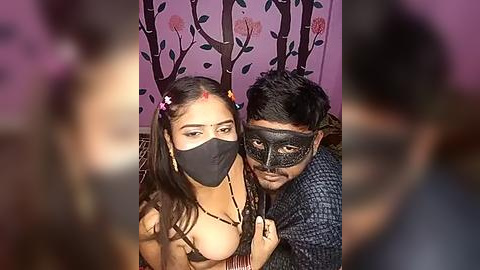 Media: Video of two people with dark skin, one with long braided hair, wearing a black face mask and a flower headpiece, the other with a black face mask, dark hair, and a patterned shirt, against a purple wall with tree and flower patterns.