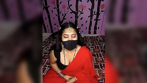 Media: Video of a woman in traditional Indian attire, wearing a red sari with a black mask, gold jewelry, and a bindi, seated on a patterned floor, against a purple wall with black tree and leaf designs.