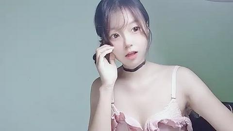 Media: A video of an East Asian woman with light skin, wearing a pink lace lingerie top, talking on a phone, against a plain light gray wall.