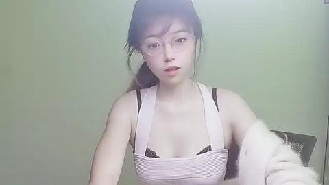 Media: Video of a young Asian woman with long black hair, wearing glasses, a white tank top, and a fluffy jacket, against a plain green background.