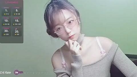 Media: Video of a young Asian woman with fair skin, wearing glasses, a beige off-shoulder sweater, and a contemplative expression, set against a plain green background.