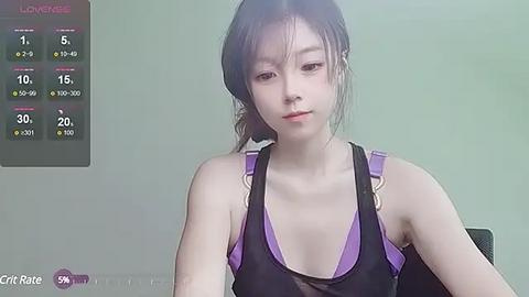 Media: Video of a young Asian woman with fair skin and black hair, wearing a black tank top, seated in an indoor setting with a digital weather display.