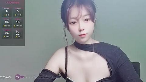 Media: Video of a young East Asian woman with pale skin, dark hair tied back, wearing a black tank top with a cutout revealing her left breast. Background shows a green wall and a screen displaying a \"Love Meter\" with stats.