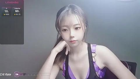 Media: Video of an East Asian woman with pale skin, straight black hair, and a slender build, wearing a purple halter top. She sits with one hand on her cheek, in a dimly lit room with a gray background.