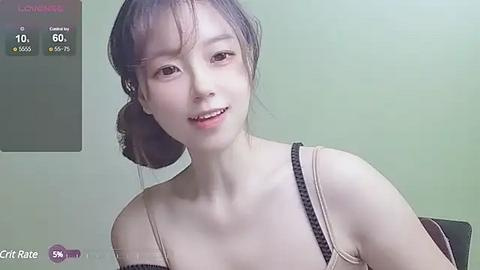 Media: A video of an East Asian woman with light skin, brown hair in a bun, wearing a beige camisole, smiling softly. Background shows a green wall with a weather app displaying 10\u00b0C.